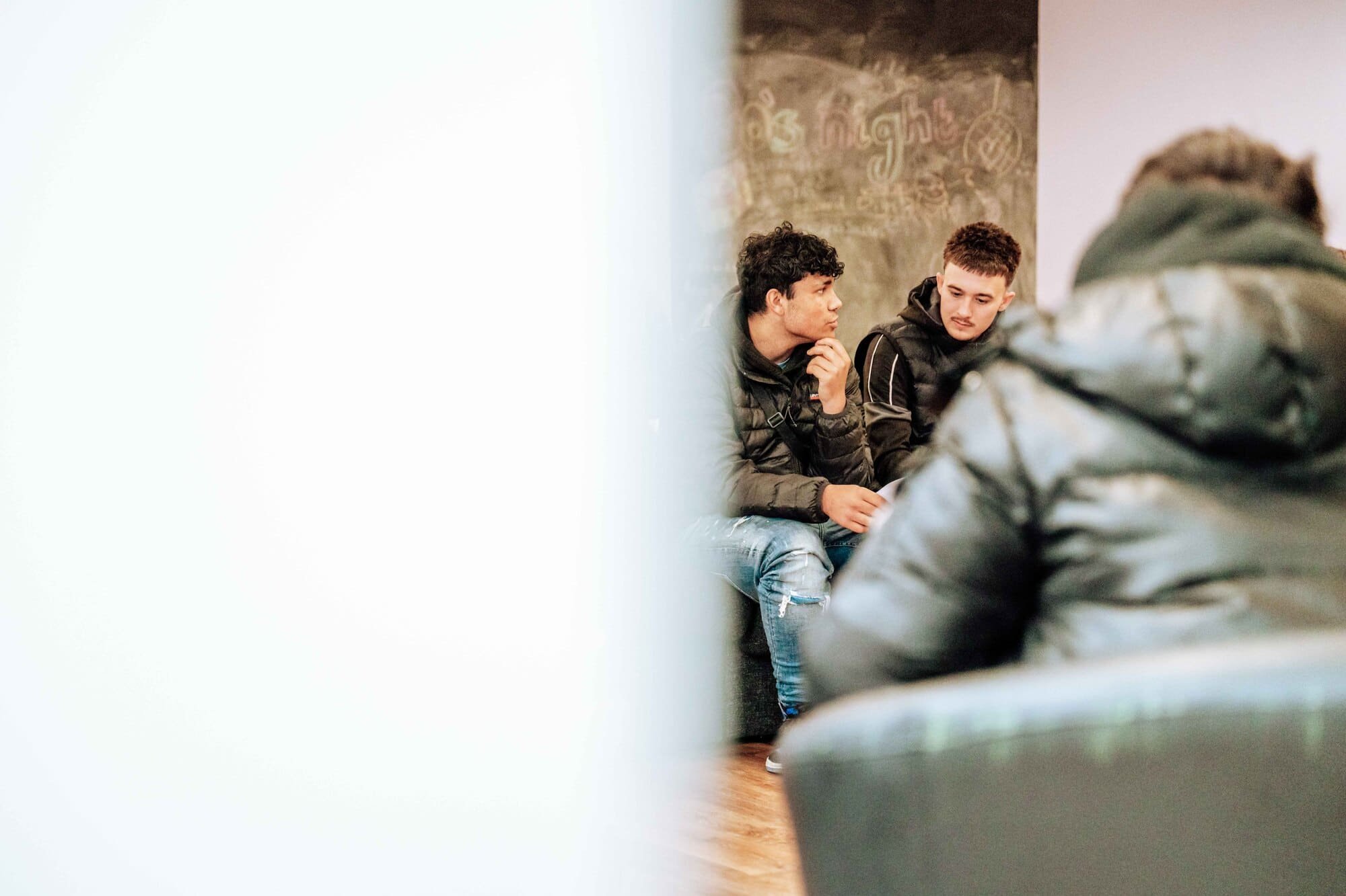 Two young men sitting and talking in a casual setting, with one of them thinking or pondering and the other looking at him, partially obscured in the foreground.