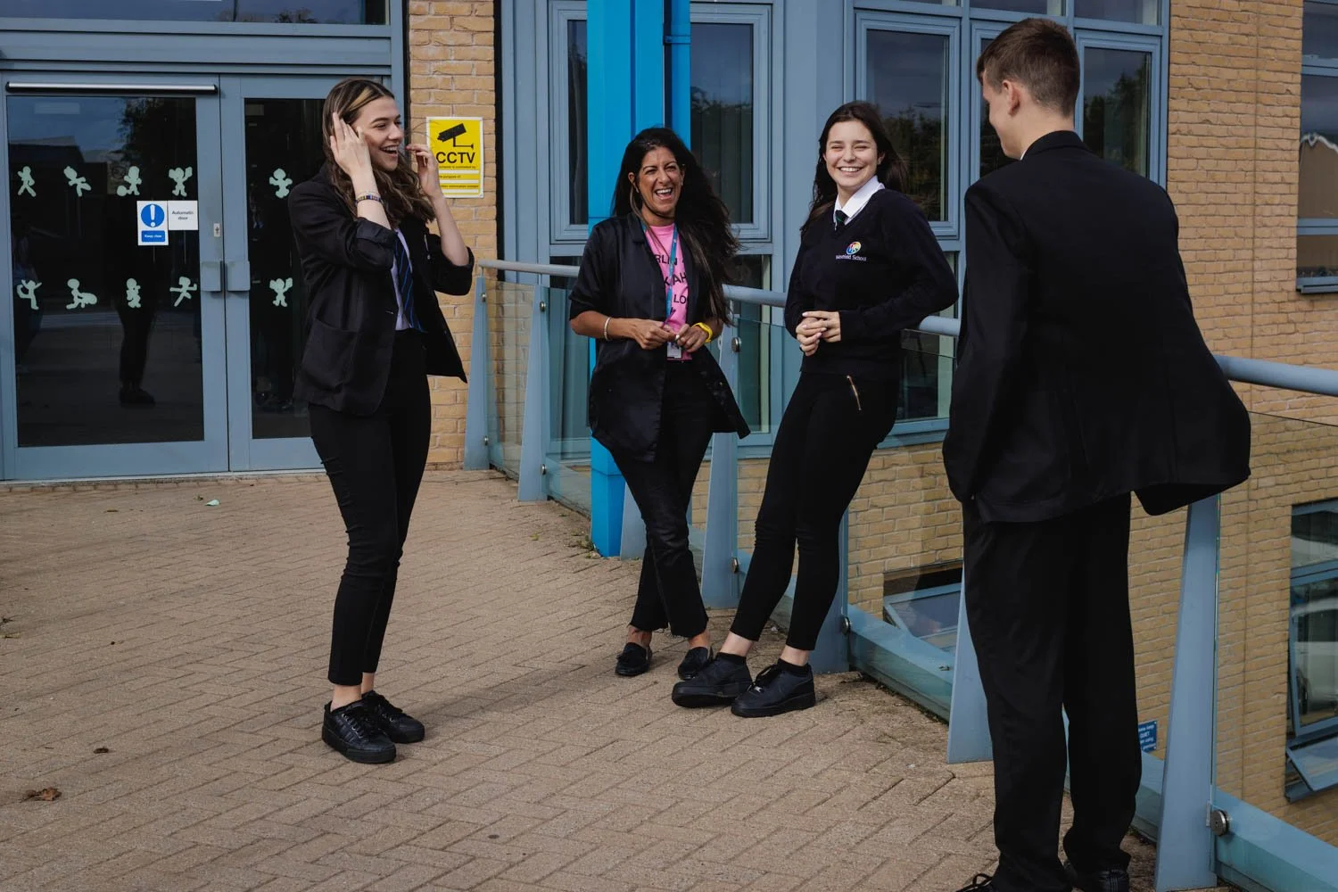 Four students, three women and one man, are standing outside a school building and laughing together. They are wearing school uniforms and appear to be having a conversation.