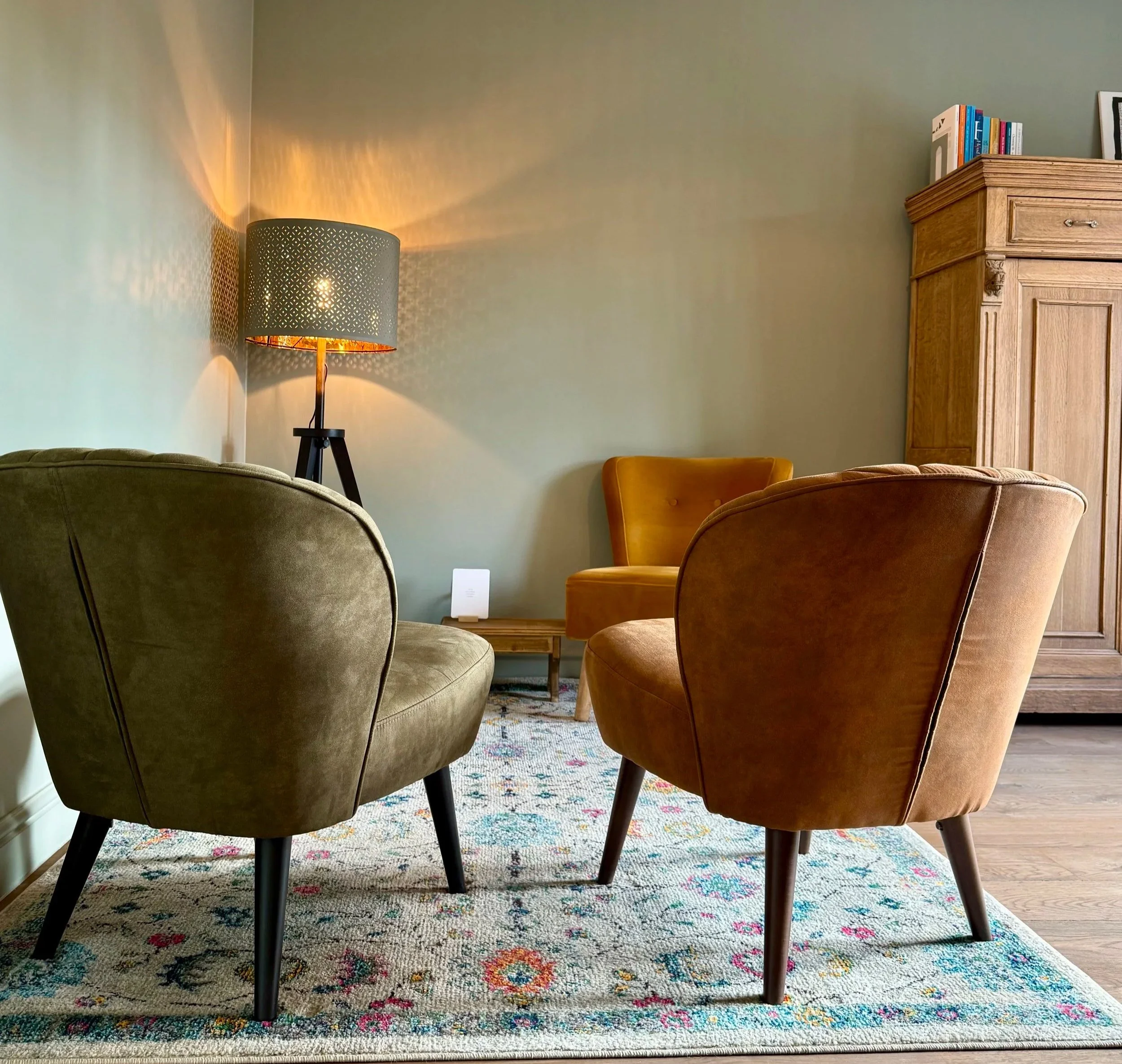 Een zithoek met drie vintage-stoelen in oranjerood, mosterdgeel en olijfgroen, een houten salontafel, een vloerlamp met een gemêleerd donkerblauw lampenkap, een houten kast met boeken, en een geruite tapijttegel.