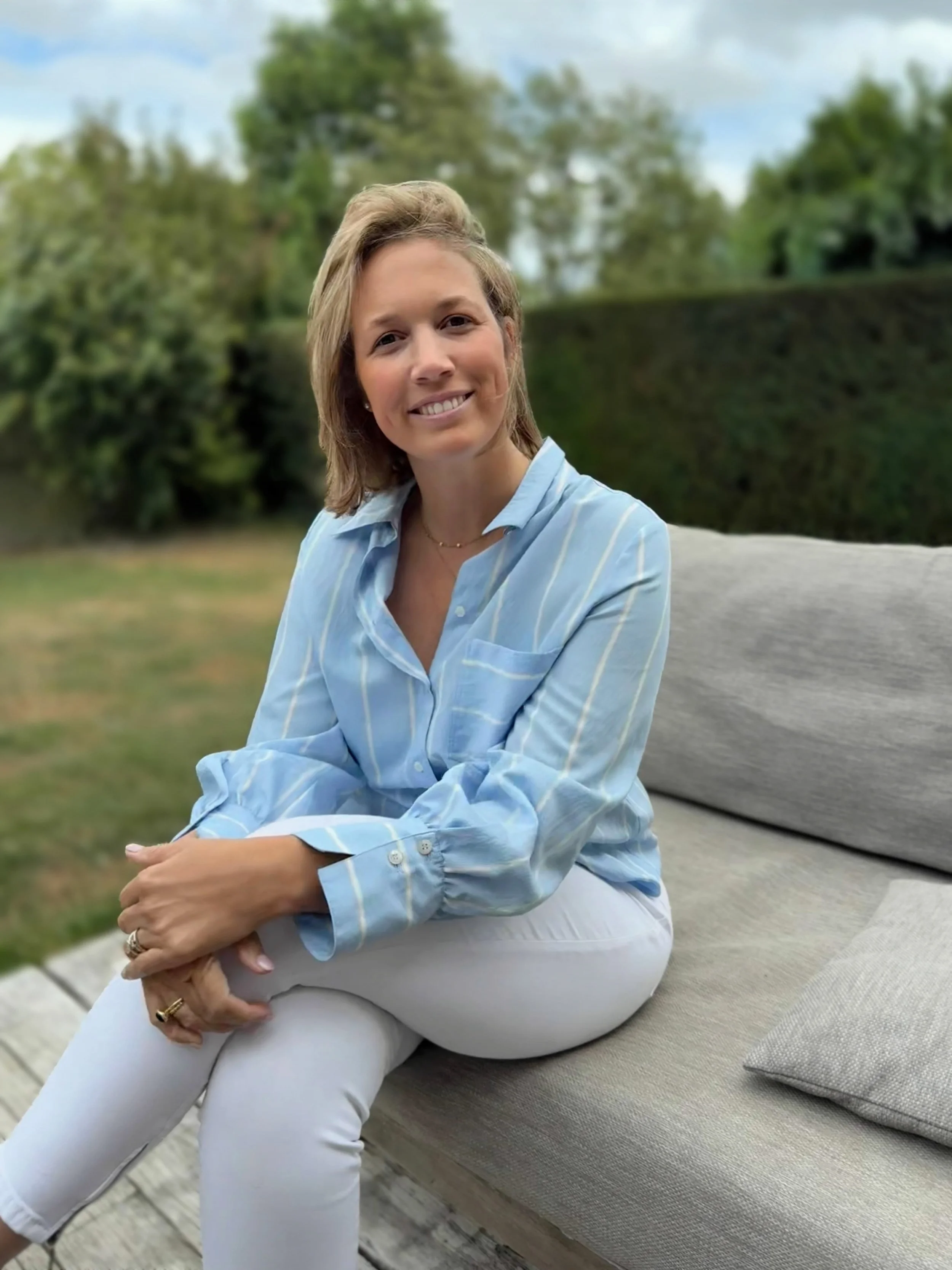 Een lachende vrouw met lichtbruin haar zit op een bank buiten, omgeven door groen en bomen.