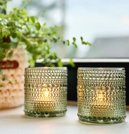 Twee kaarsen in glazen houders met een bubbeltjespatroon op een tafel, met planten op de achtergrond.