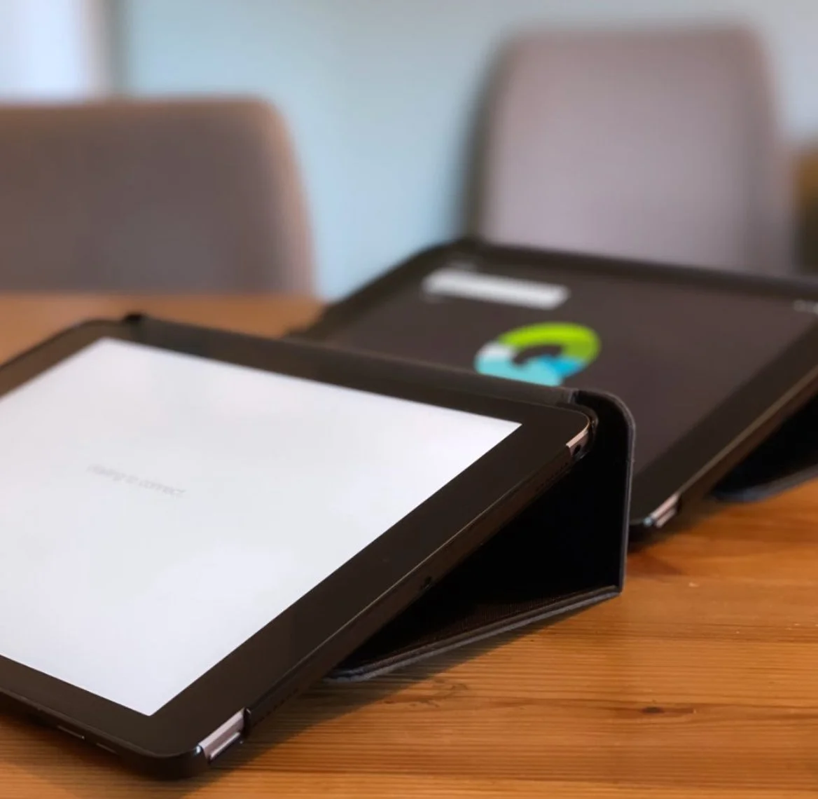 Een tablet in een hoesje ligt op een houten tafel, met een tweede apparaat op de achtergrond.