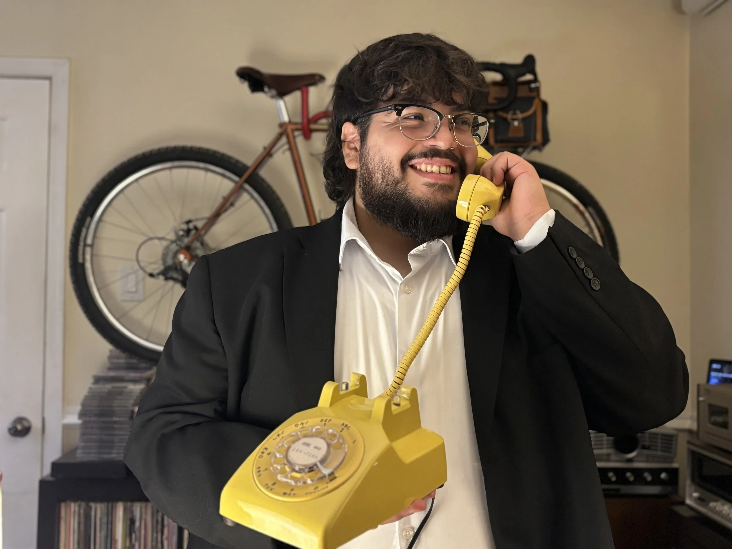 A man with glasses and a beard, wearing a black suit and white shirt, smiling while talking on a yellow rotary phone.