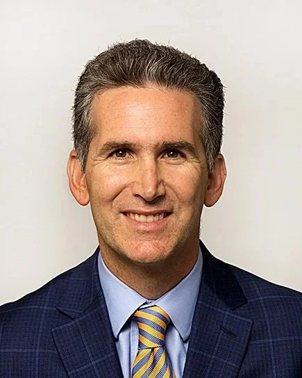 A professional man with short, gray hair, dressed in a navy blue suit, light blue shirt, and a yellow striped tie, smiling at the camera.