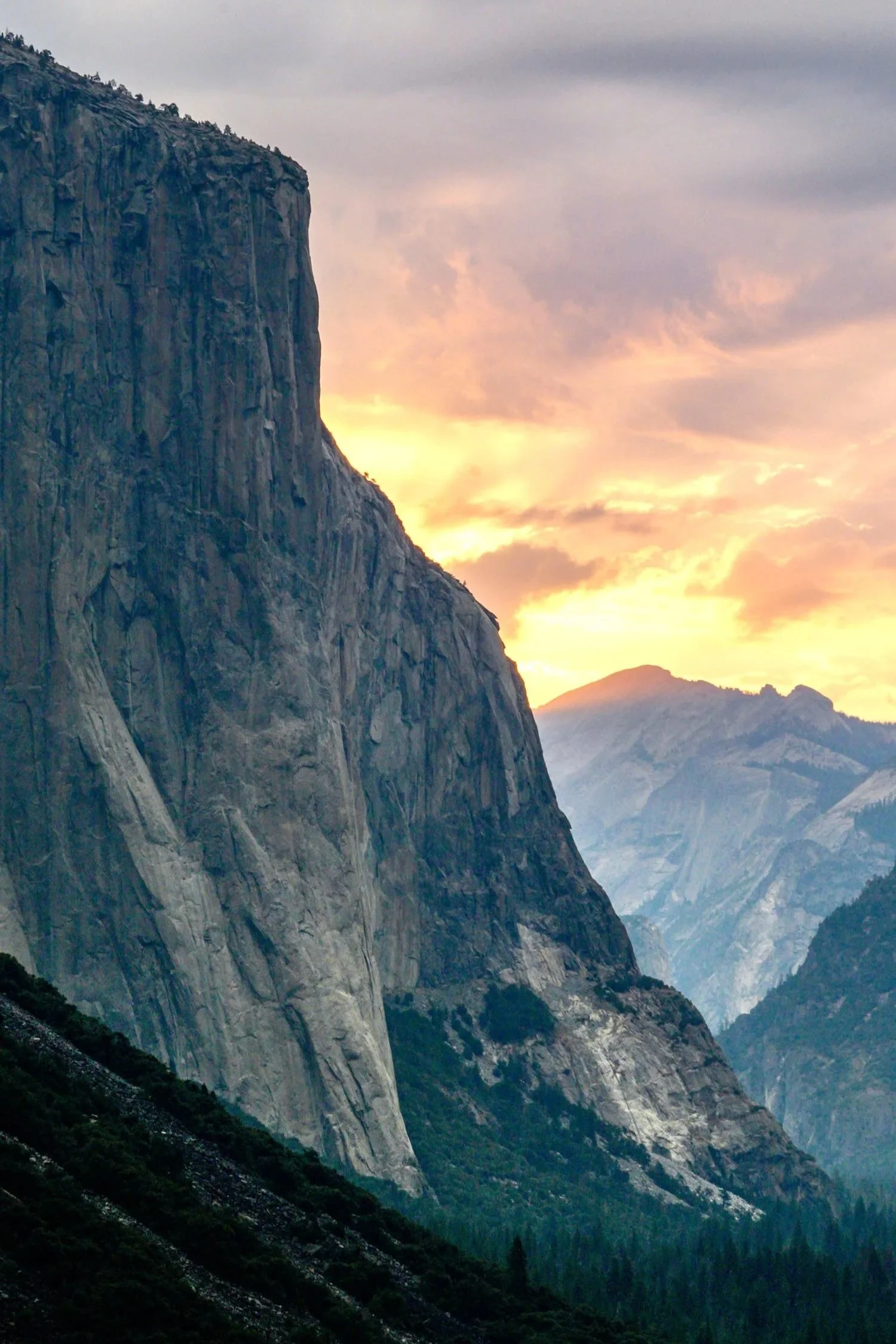 A scenic view of a mountain landscape at sunset, featuring a prominent granite cliff and distant mountain ranges under a partly cloudy sky.