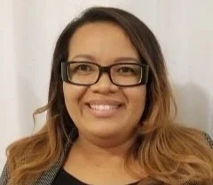 A woman with long hair wearing glasses and a black outfit, smiling at the camera.
