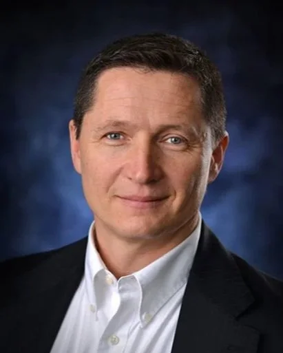 Professional headshot of a man in a white shirt and dark blazer against a dark, textured background.