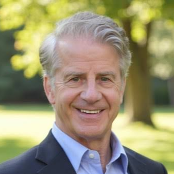 Headshot of a smiling middle-aged man with gray hair, wearing a blue shirt and dark blazer outdoors with trees in the background.