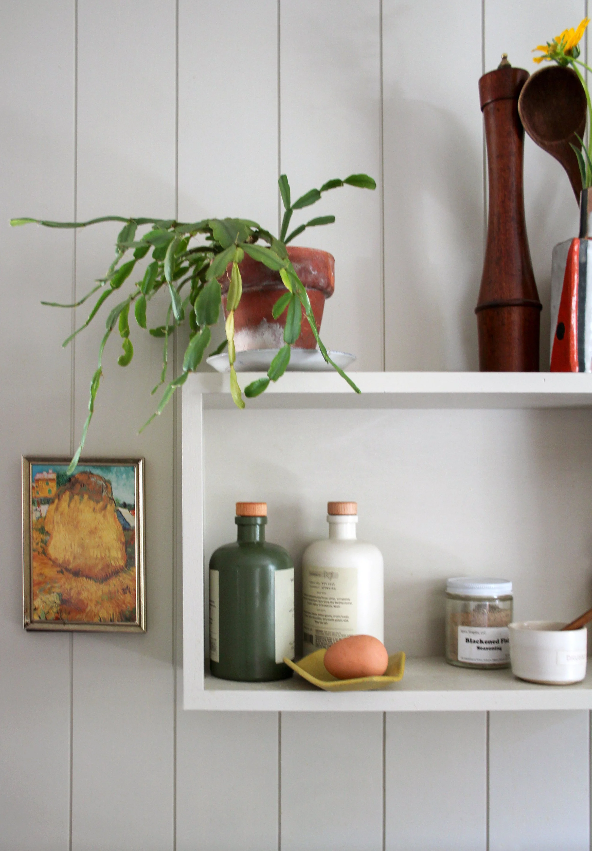 A white wall shelf with potted plant, decorative wooden objects, bottles, an egg, a jar, and a bowl. A small framed picture is on the wall nearby.