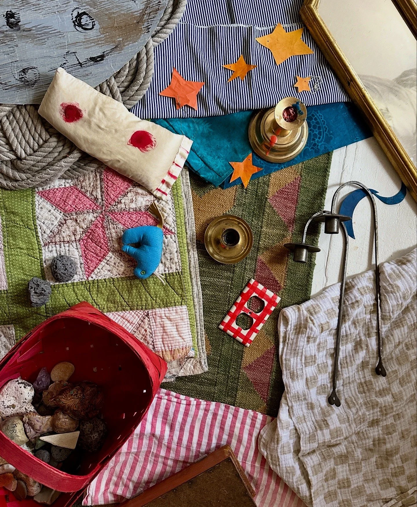 A collection of various objects on a table or surface, including a wrapped pillow, star-shaped paper decorations, brass candle holders, a small red and white checkered box, a small red basket filled with stones or crystals, a toy blue elephant, a glass of dark liquid, patterned fabric, and a striped cloth.