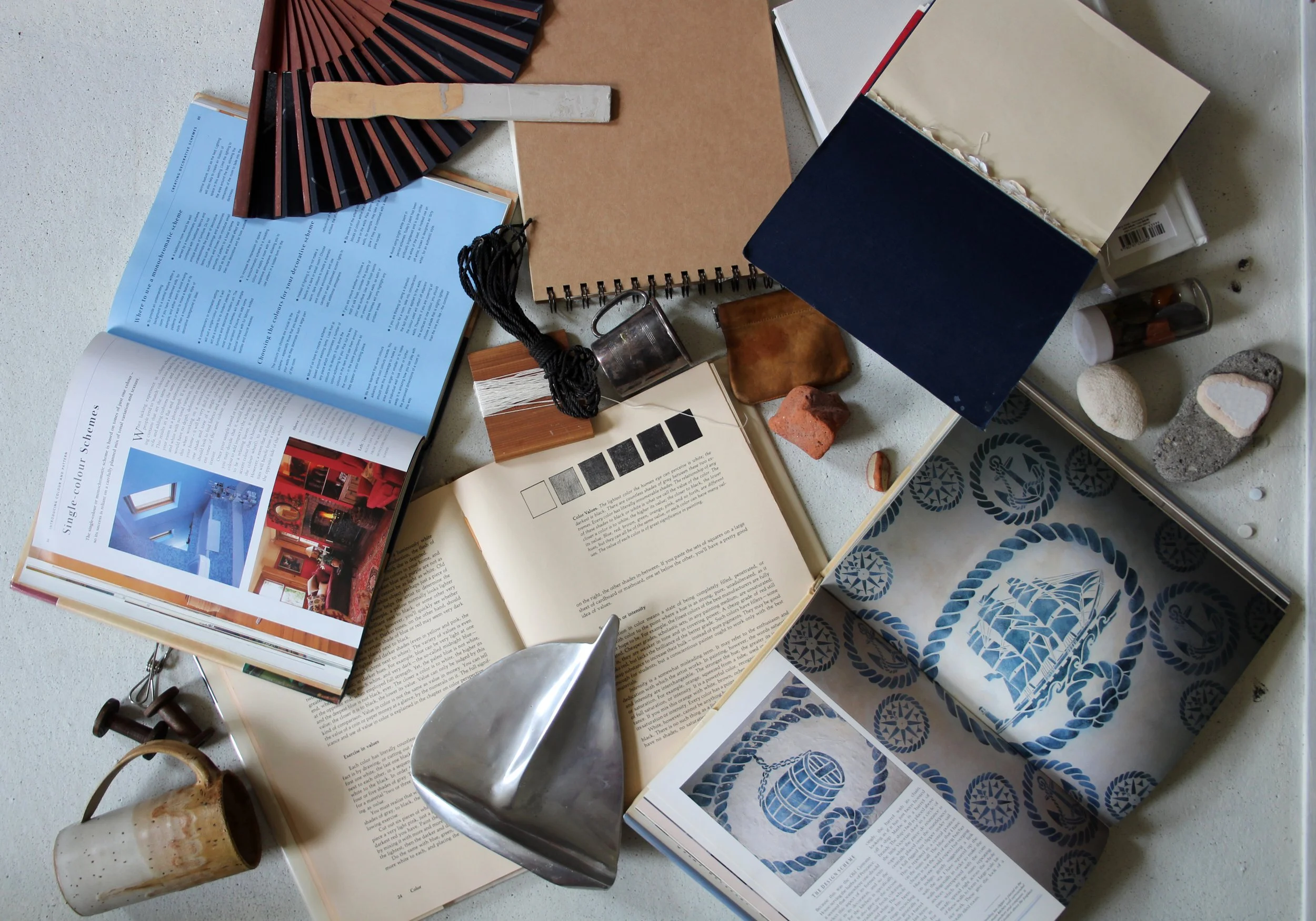 A variety of interior design and decorating items spread out on a white surface, including open books and magazines, fabric samples, small rocks and stones, a ceramic mug, a metal scoop, a spiral notebook, a box with patterned paper, a fan, and other small objects.