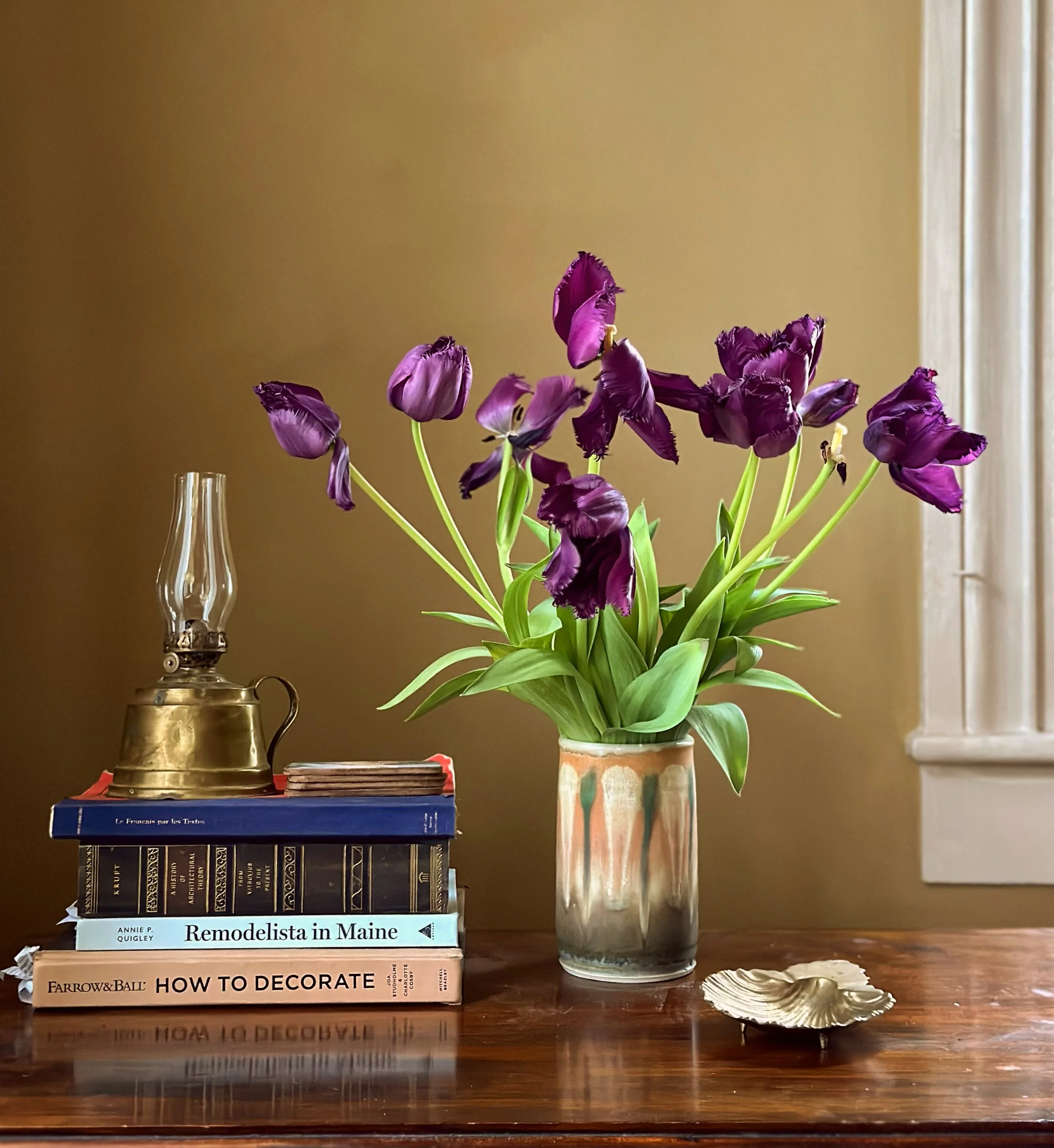 A still life composition on a wooden table with a stack of books including 'Remodelista in Maine' and 'How to Decorate,' a vintage oil lantern, a ceramic vase with purple tulips, and a gold shell ornament, set against a beige wall with white window trim.