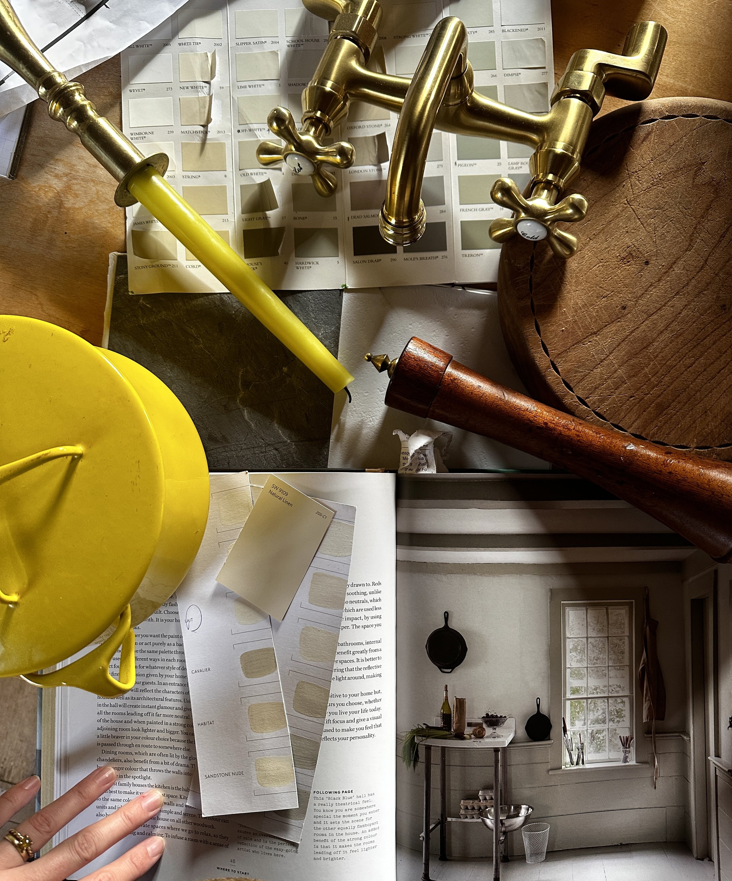 Arrangement of interior design samples, including paint color swatches, metallic and wooden faucet fixtures, a yellow lamp, a wooden serving tray, and a printed interior decor book page, on a wooden table.