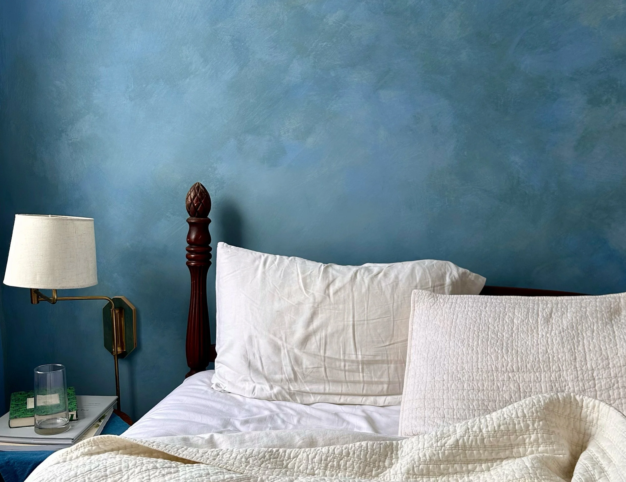 A bedroom with a bed against a blue textured wall, featuring a wooden headboard post, a white pillow, a quilt, a bedside table with a glass of water, books, and a lamp.