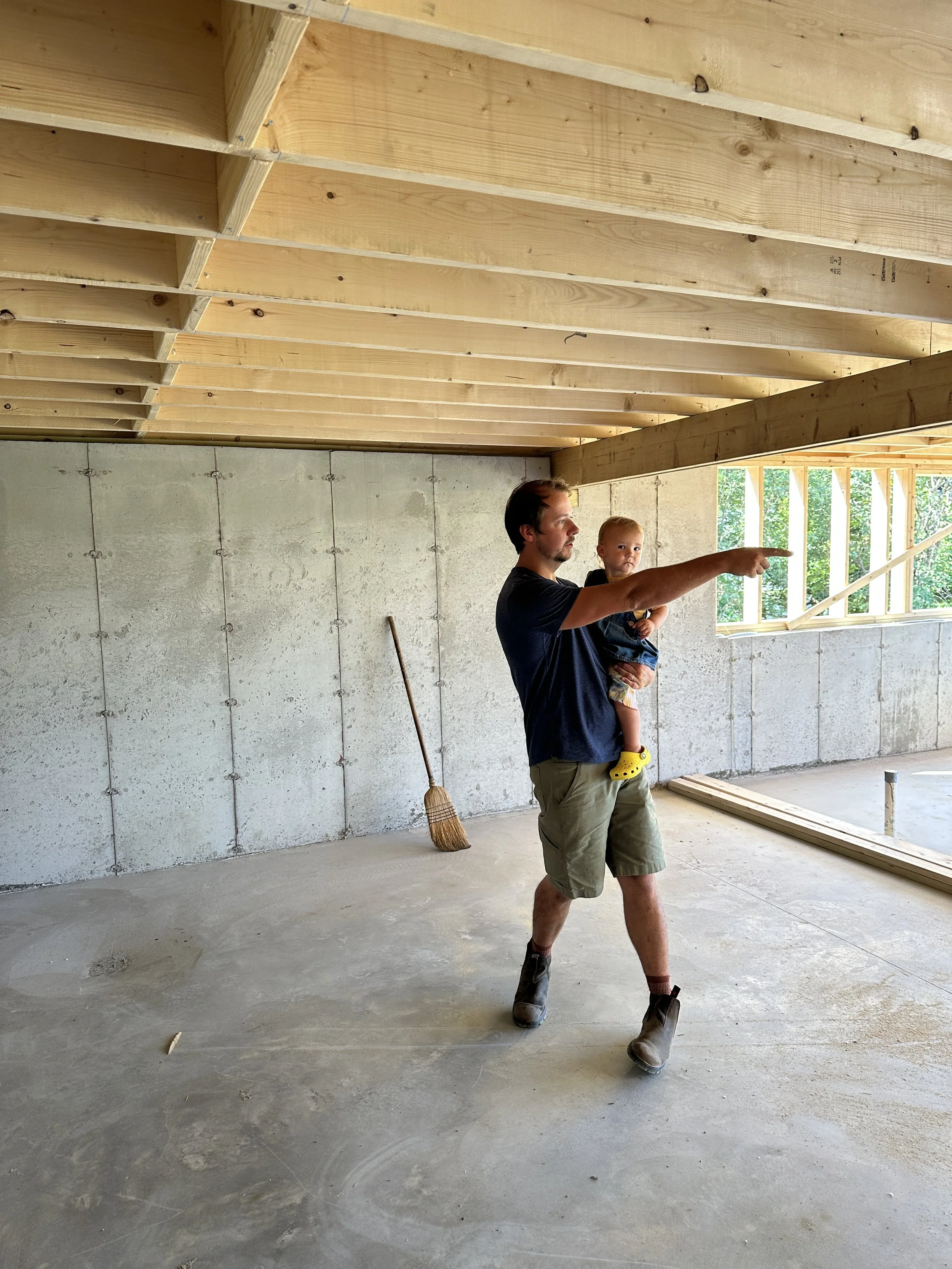 A man holding a young child in an under-construction building, pointing towards the ceiling where new wooden beams are being installed, with a broom and construction materials around.
