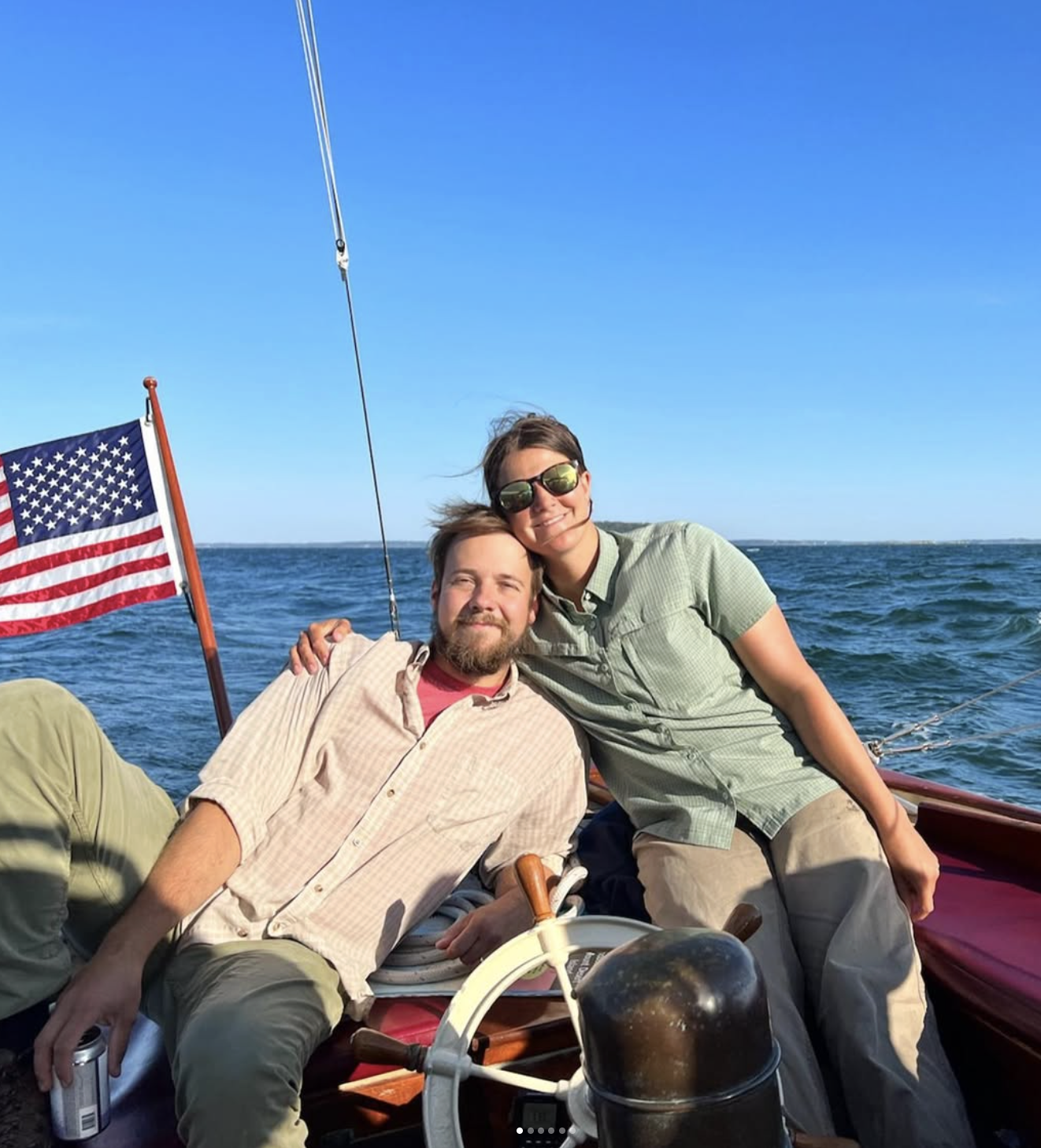 A man and woman enjoying a boat ride on the water with an American flag in the background, smiling at the camera.