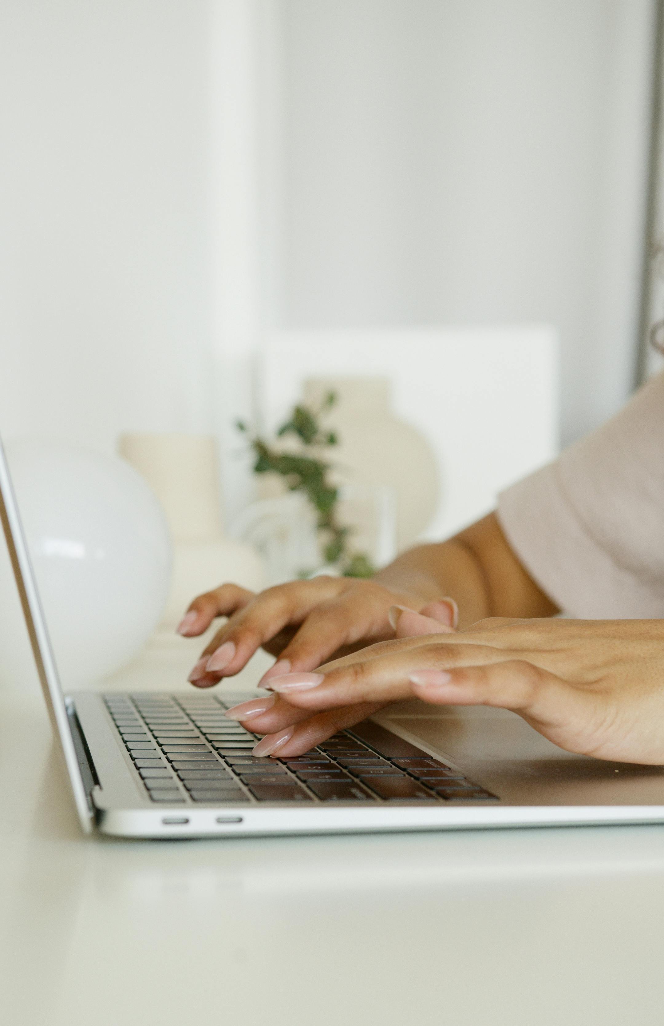 Close-up of hands typing on a silver laptop keyboard, with a white vase and a plant on a white table in the blurred background.