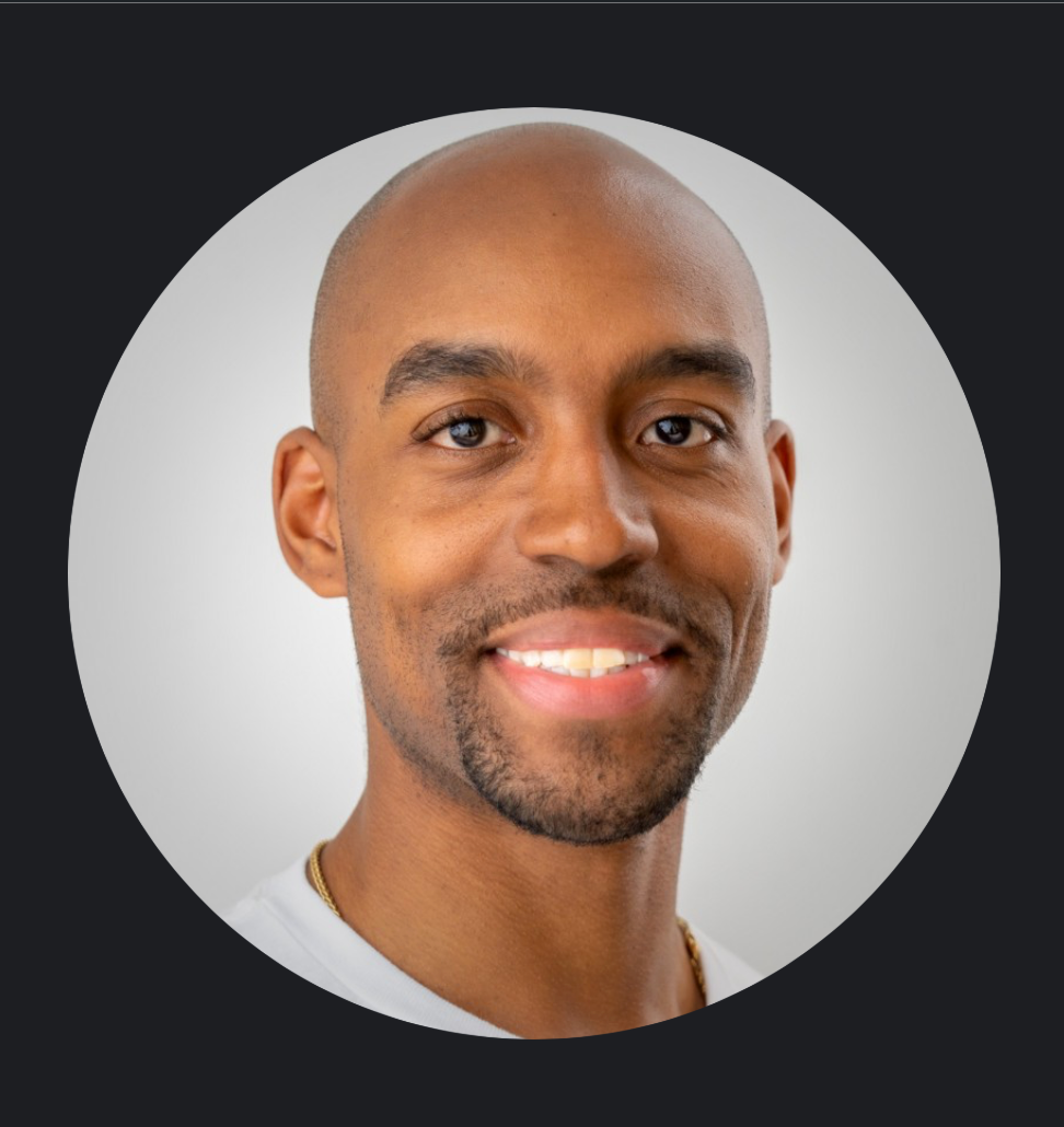 Close-up of a smiling man with a bald head, facial hair, and wearing a white shirt, against a neutral background.