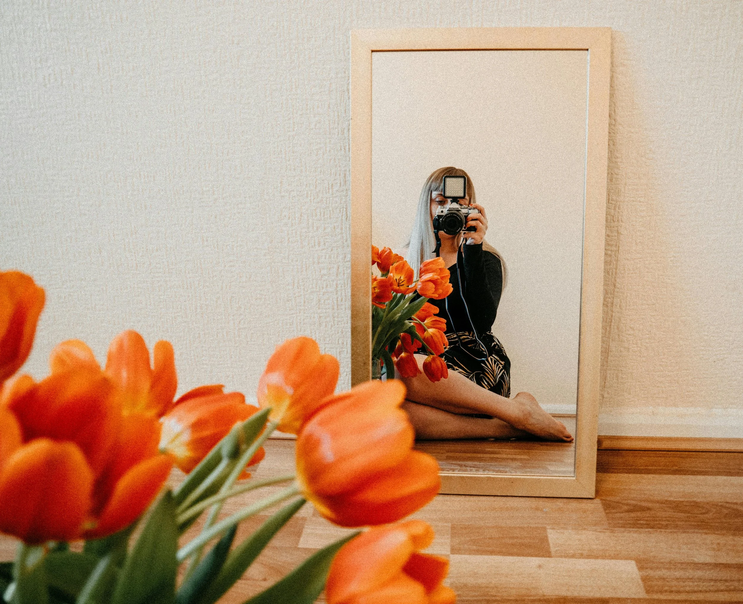 A woman taking a photo of herself in a mirror with a camera, sitting on the floor with orange flowers in the foreground.