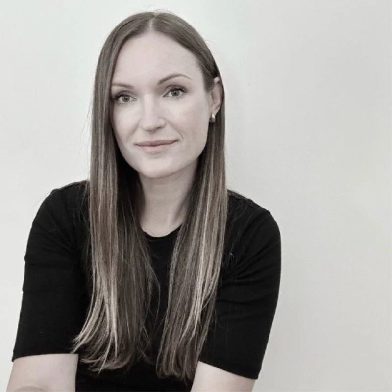 A woman with long, straight hair wearing a black shirt, smiling softly while sitting against a plain, light-colored wall.