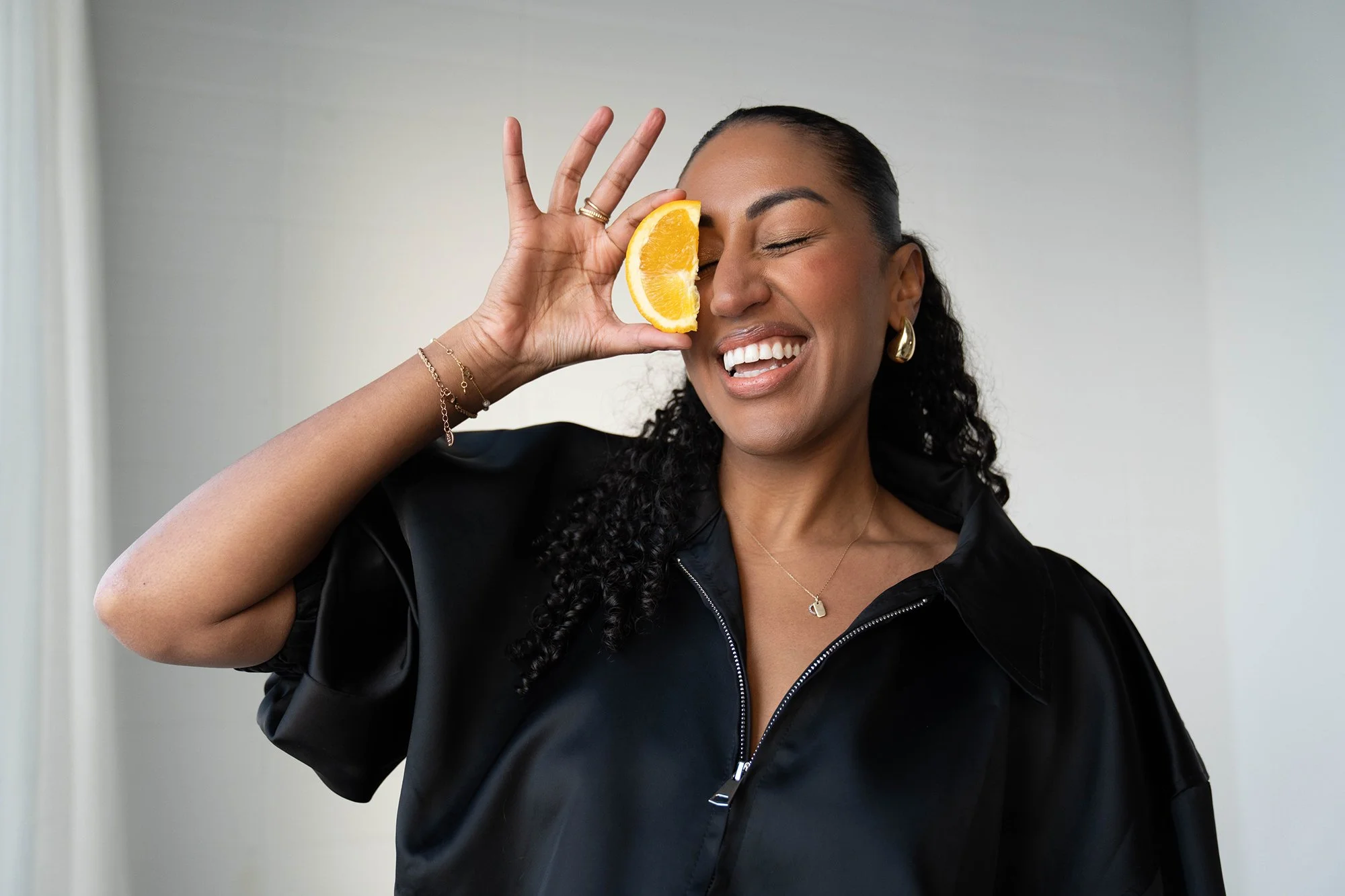 Woman with curly hair and earrings holding a lemon wedge to her eye, smiling.