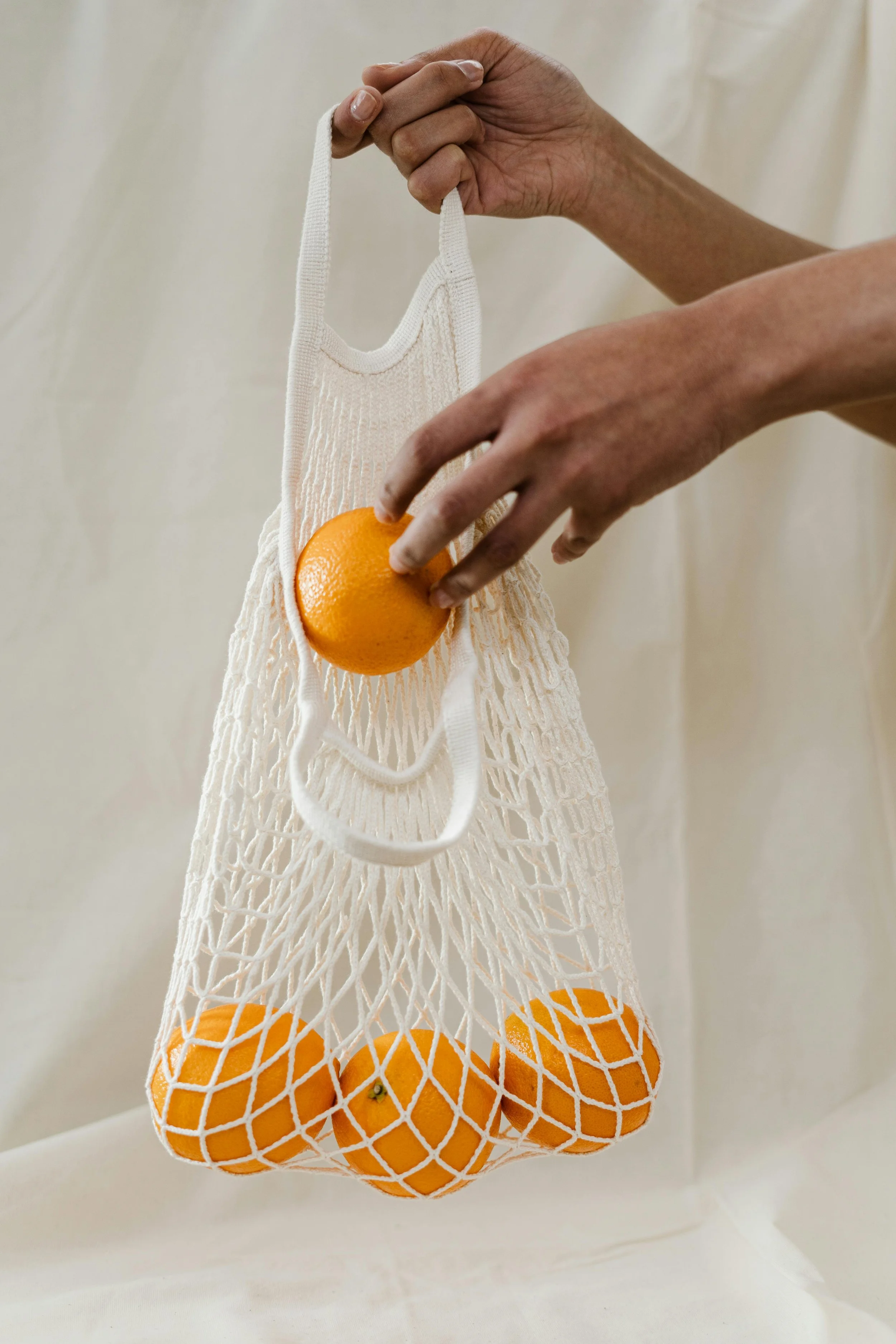 Person holding a reusable mesh bag with oranges and one orange in their hand.