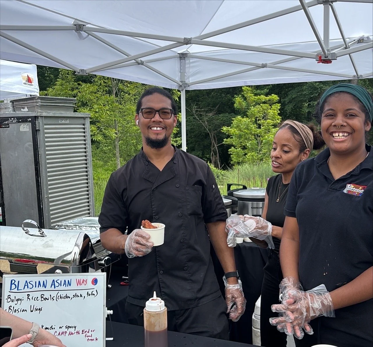 Smiling chef wearing a black chef's coat and gloves holding a small cup of food at an outdoor booth, with team members preparing items behind him under a white canopy.