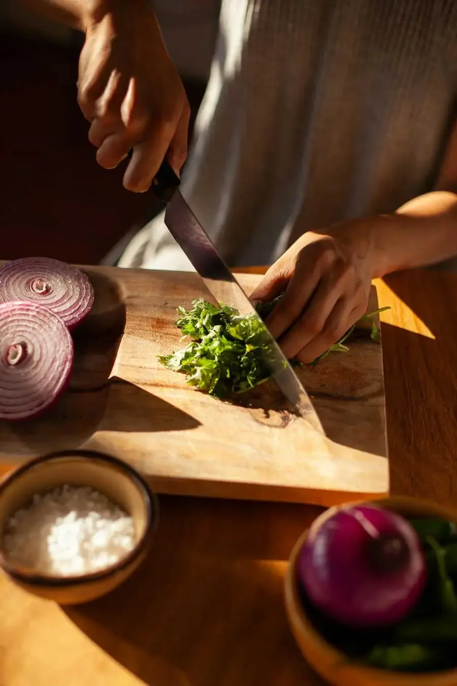 Person chopping fresh cilantro on a wooden cutting board, with sliced red onions and small bowls of salt and additional vegetables nearby.