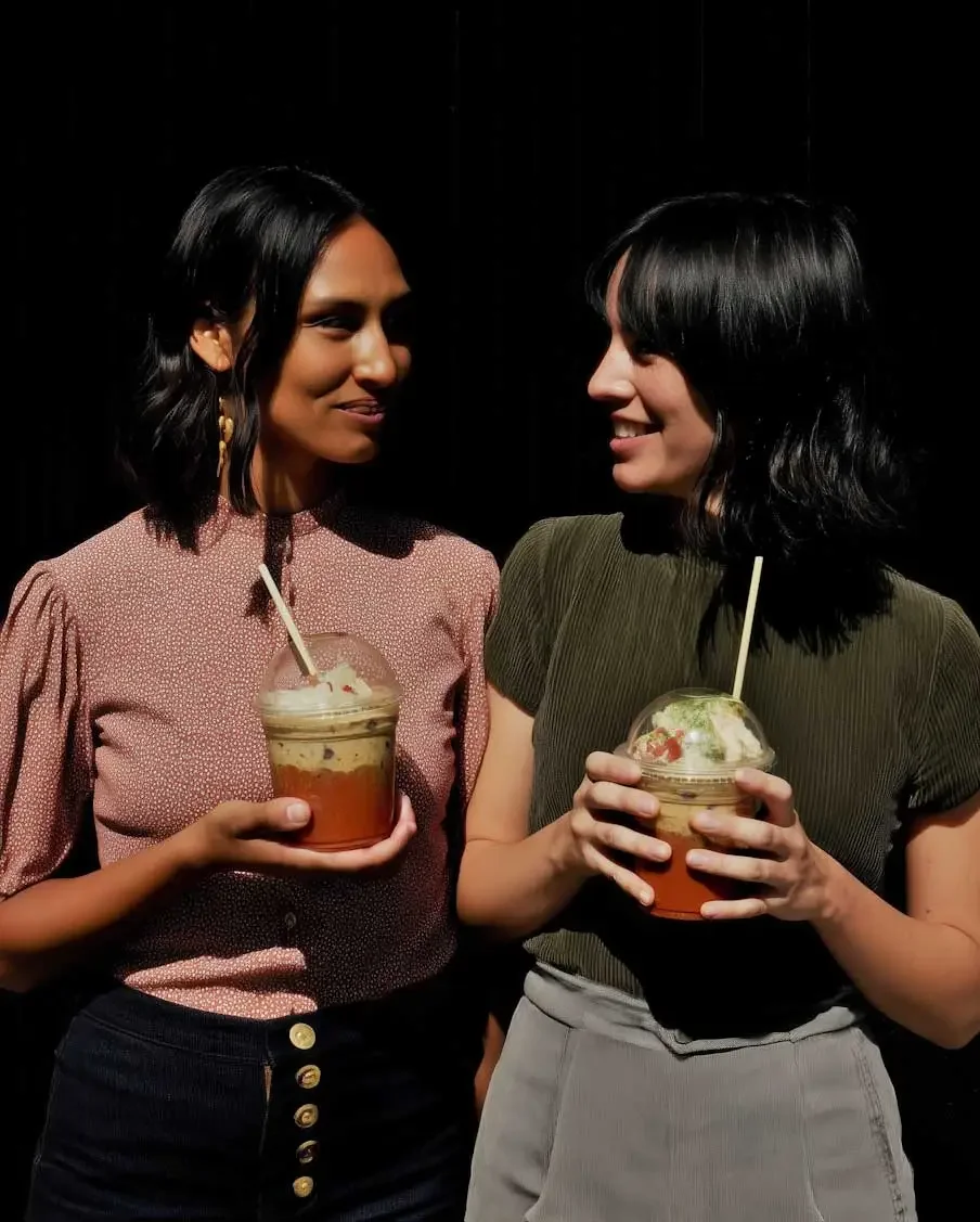 Two women holding blended frozen Matcha drinks and smiling at each other against a black background.