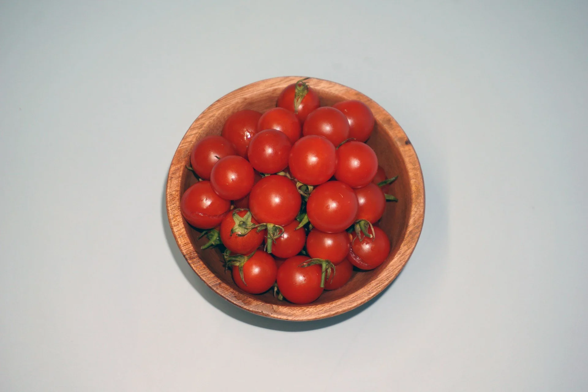  Top-down view of a wooden bowl filled with fresh red cherry tomatoes against a light background. 
