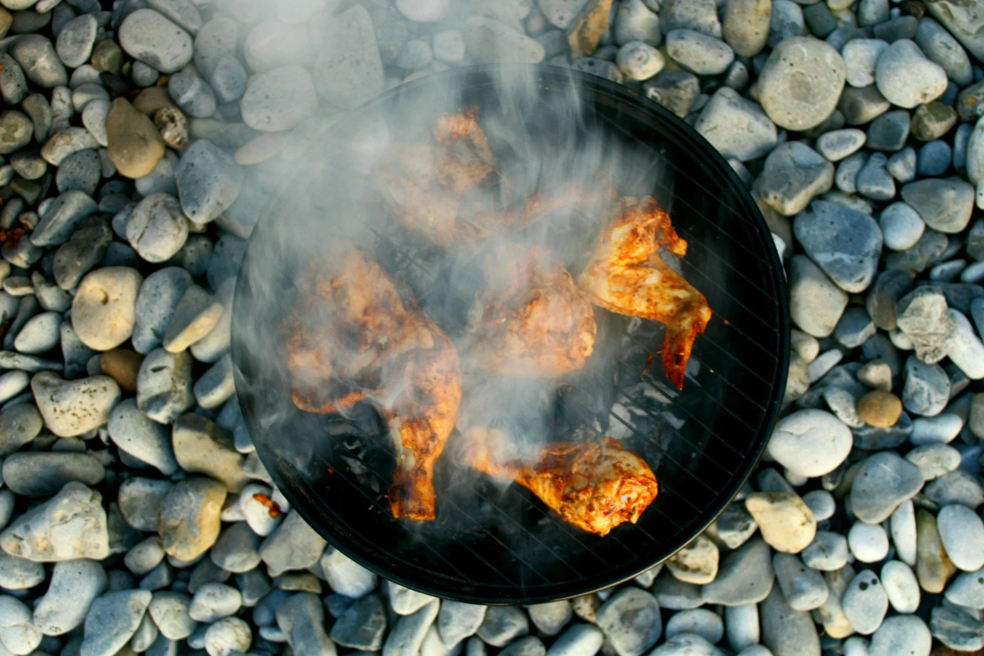  Top-down view of seasoned chicken pieces sizzling on a charcoal grill, with smoke rising over the meat and smooth gray stones surrounding the grill. 