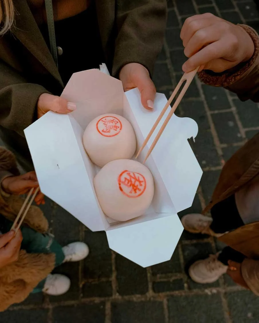 Two people holding a white box with two steamed buns with red stamps on top, using chopsticks.