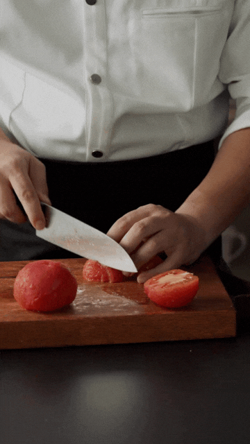  Chef in a white coat slicing peeled tomatoes on a wooden cutting board, holding a large kitchen knife with focus on the hands and ingredients. 