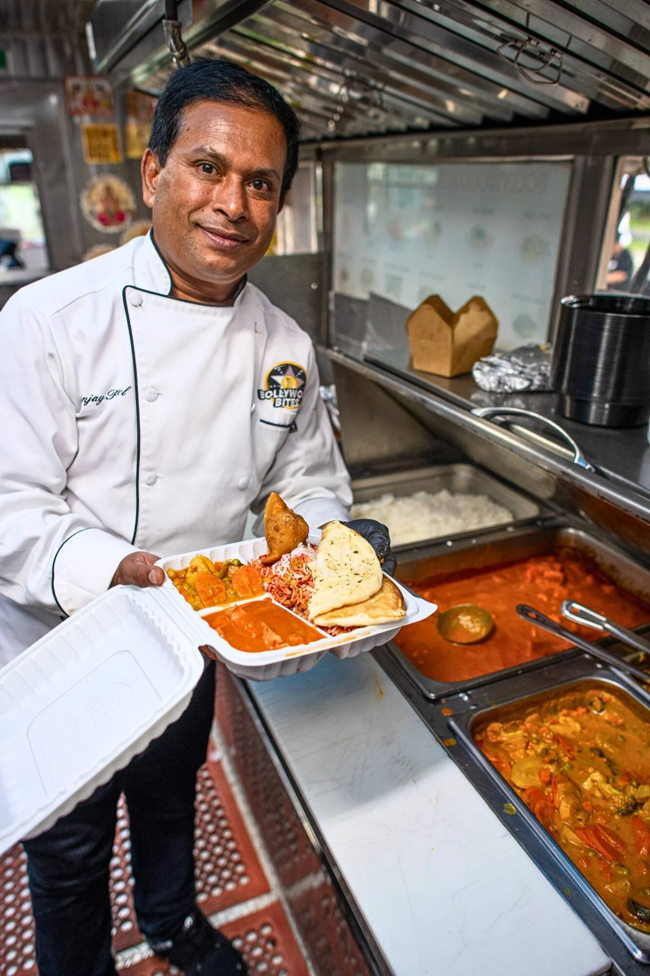 Chef Sanjay Patel of inside the Bollywood Bites food truck holding an open takeout container filled with Indian dishes, including curry, rice, samosa, and naan, with steaming trays of food visible behind him.