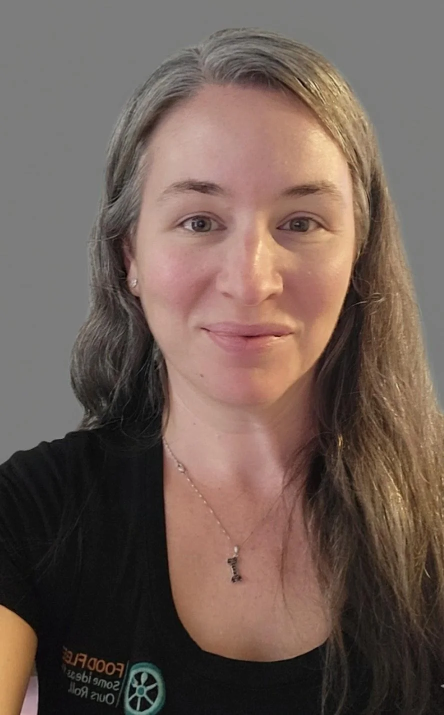 Portrait of a woman with long gray-streaked hair, wearing a black top and a delicate necklace, looking directly at the camera against a neutral background.