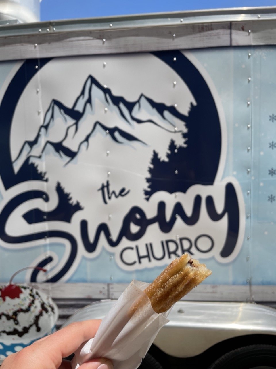Hand holding a churro wrapped in paper in front of a food truck decorated with a snowy mountain logo reading “The Snowy Churro.”