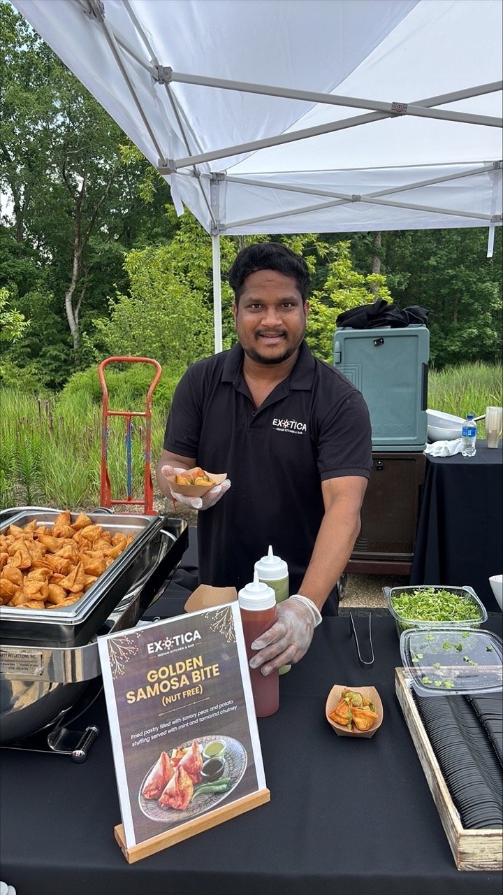 Vendor standing behind an outdoor food booth, smiling as he holds a tray of samosas next to a chafing dish filled with more; a sign reading “Golden Samosa Bite” is displayed in front of the table with sauces and toppings.