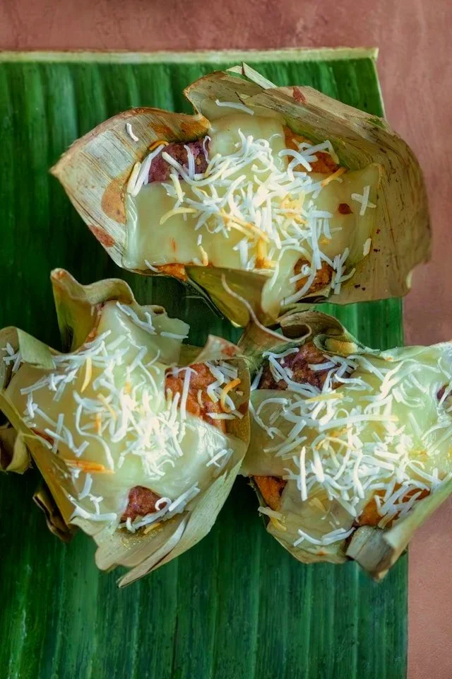 Three unwrapped tamales topped with shredded cheese on a green banana leaf.