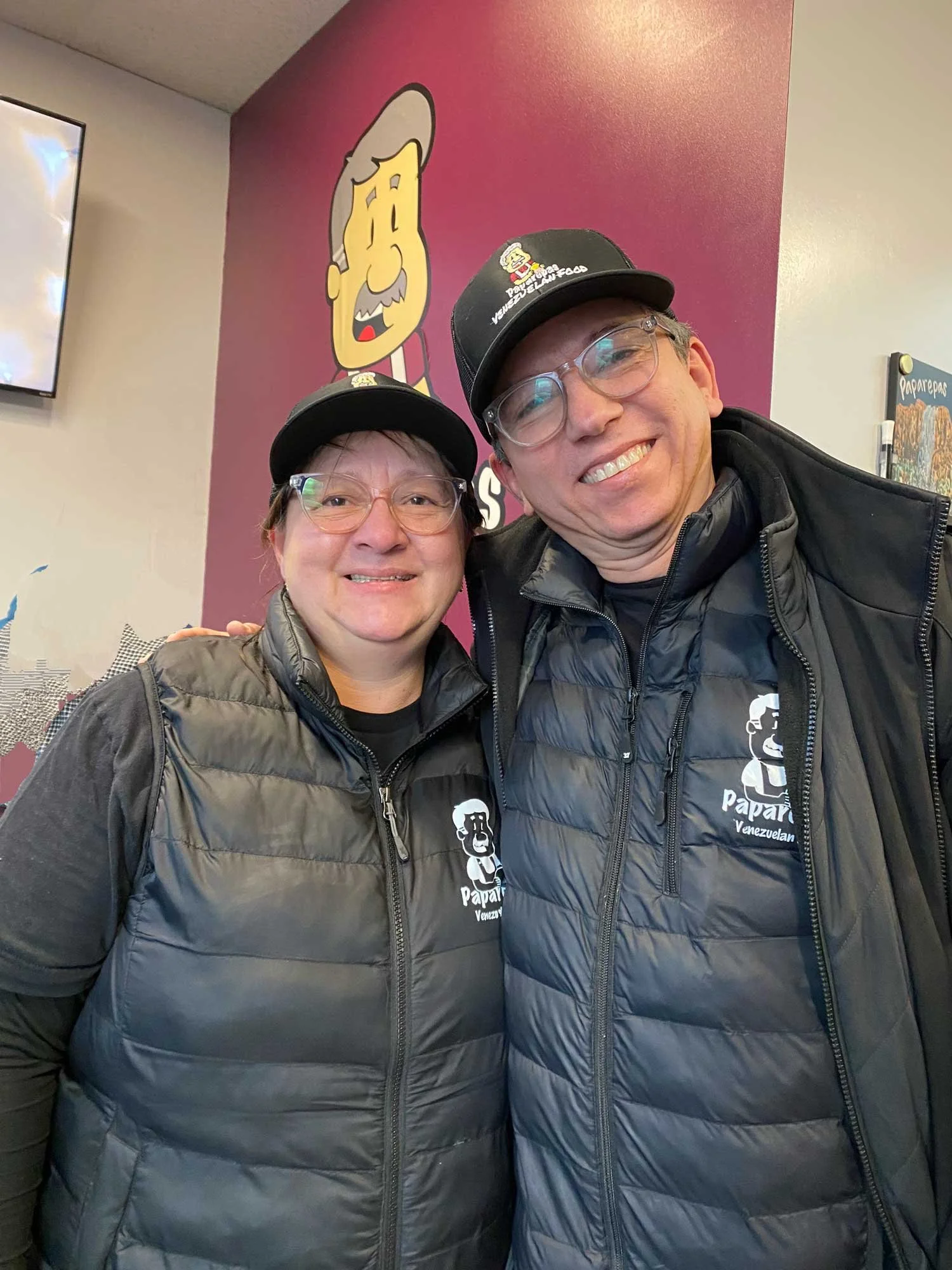Two smiling food business owners wearing matching branded caps and puffer vests, standing close together inside their restaurant or food service space.
