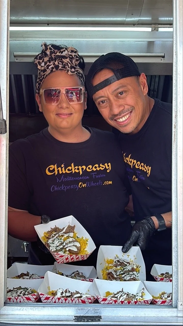 Two smiling food truck workers wearing Chickpeasy shirts, holding trays of Mediterranean fusion dishes topped with meat and sauce inside their service window.