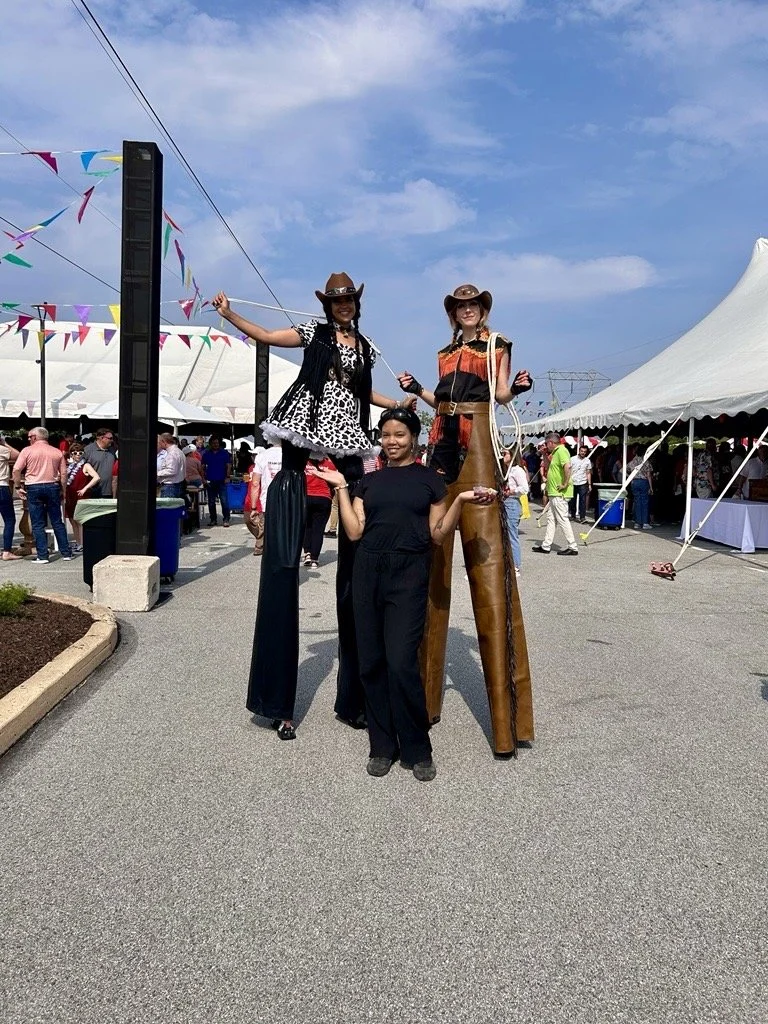 Two performers on tall stilts dressed in cowboy-themed outfits posing with a smiling woman between them at an outdoor event with tents, crowds, and festive decorations.