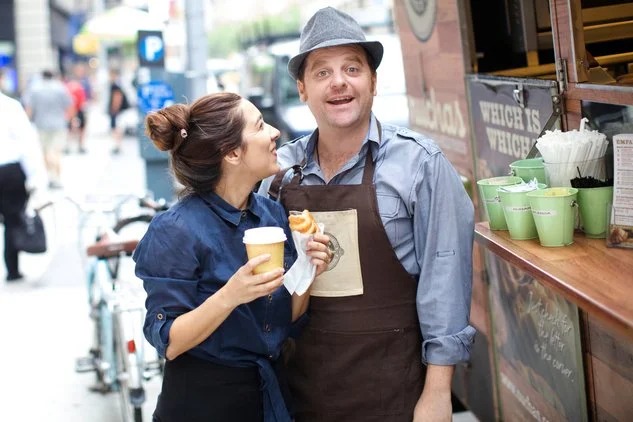Man and woman standing beside a food truck on a city street, smiling and chatting while holding coffee and a pastry, with cups and utensils displayed at the service window.
