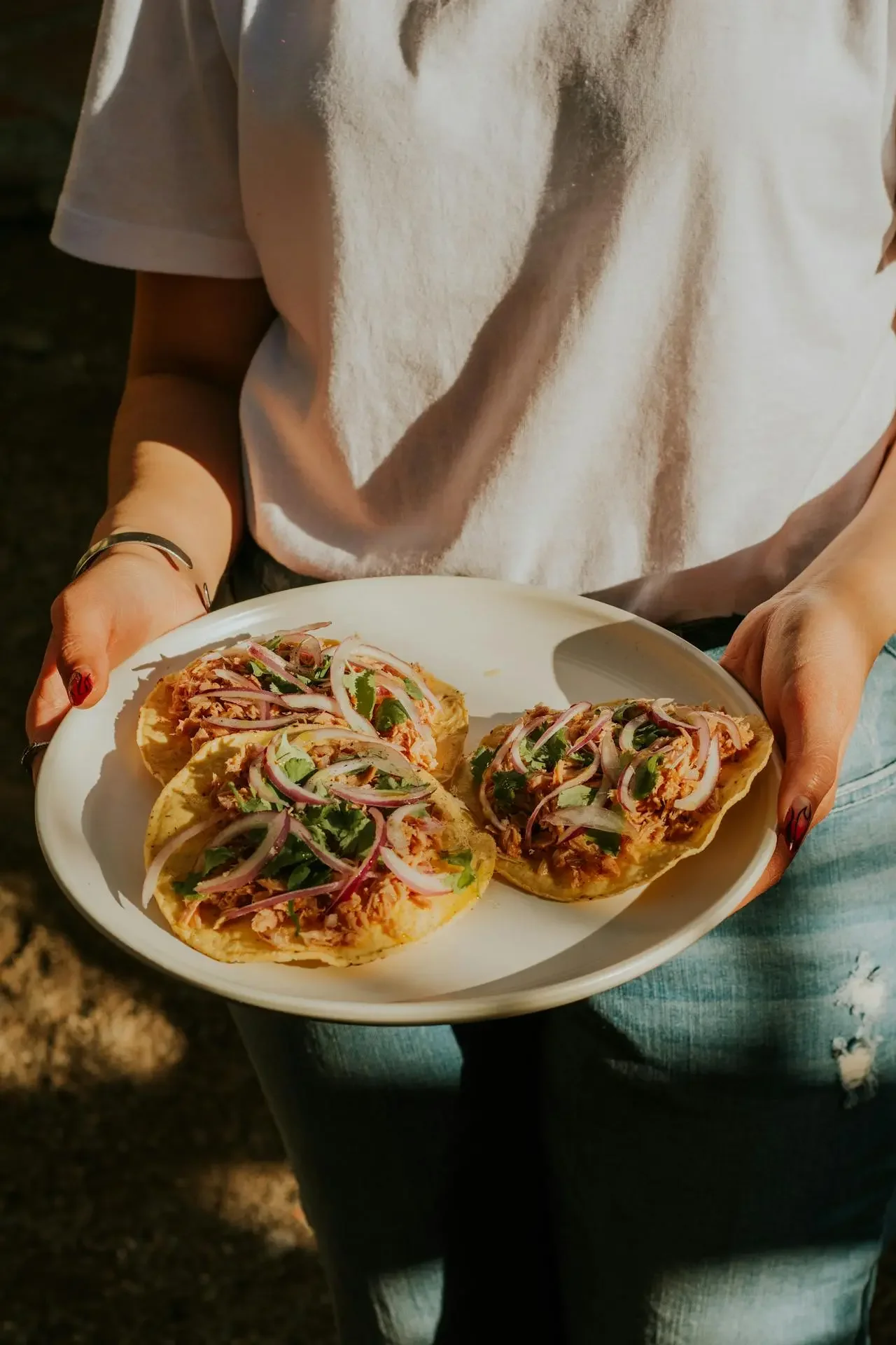 Person holding a white plate with three tacos topped with onions and cilantro