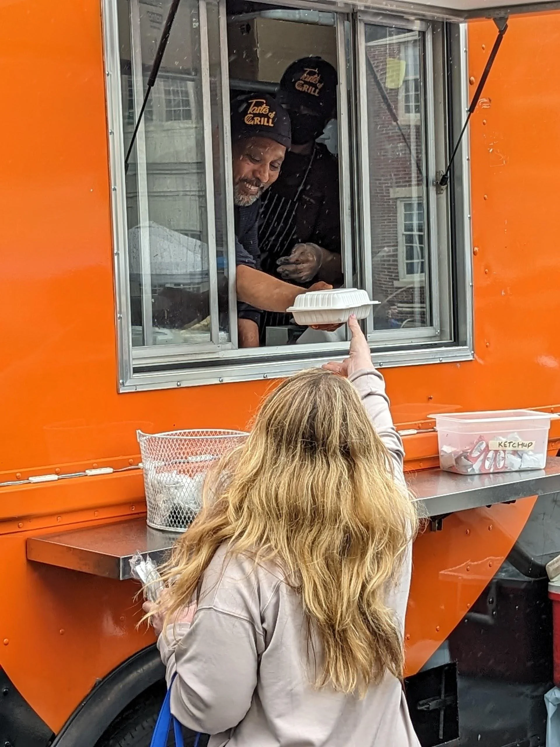Customer reaching up to receive a takeout container from a food truck worker through the service window of an orange food truck.