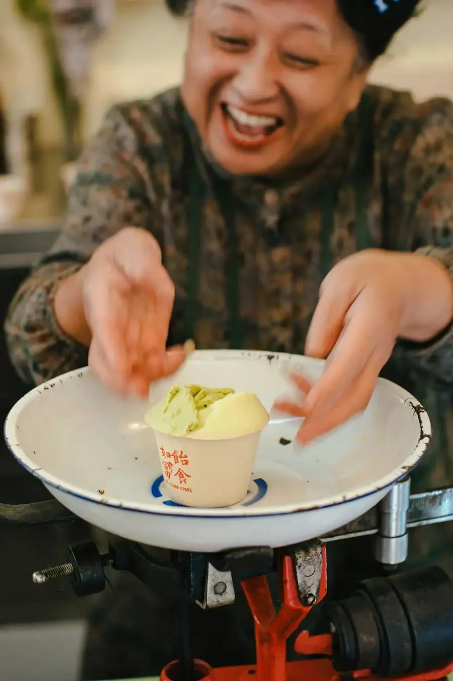 An older Asian woman with a joyful expression reaching for a cup of green ice cream on a vintage food scale, with a blurred background.
