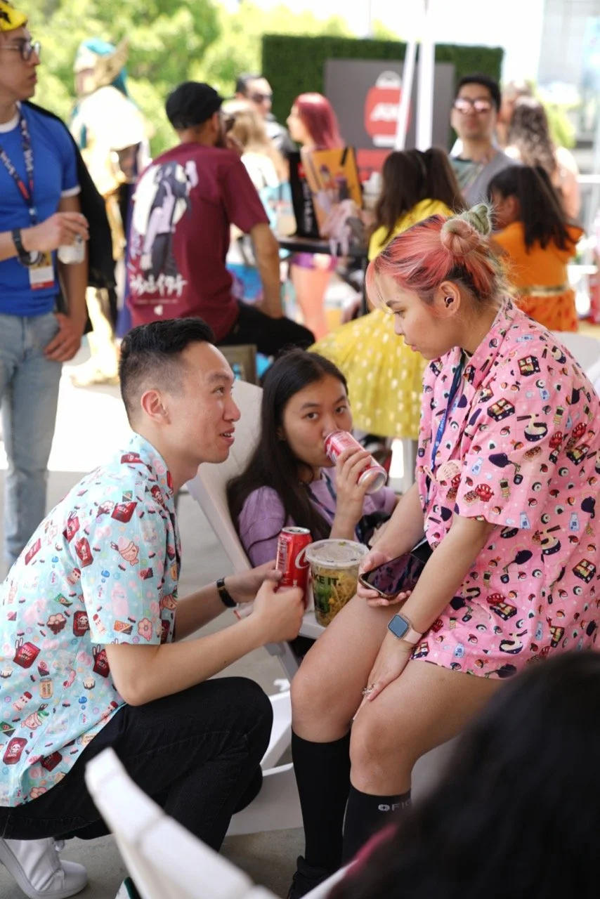 Group of young people at Animé Los Angeles, chatting and drinking sodas; two wear colorful patterned shirts while another sits with a large cup of noodles in her lap, surrounded by a busy crowd.