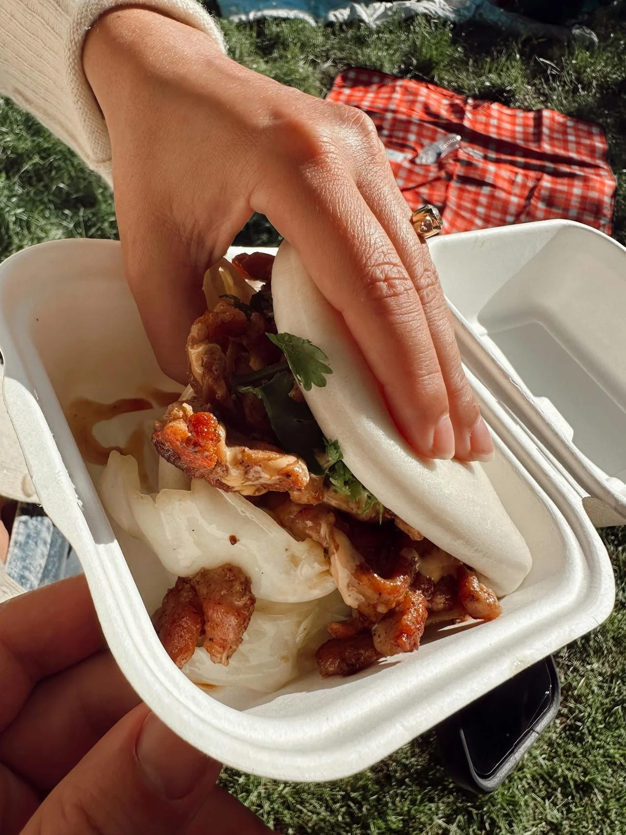 Person holding a steamed bao bun sandwich filled with meat and vegetables over a takeout container with more bun filling inside.