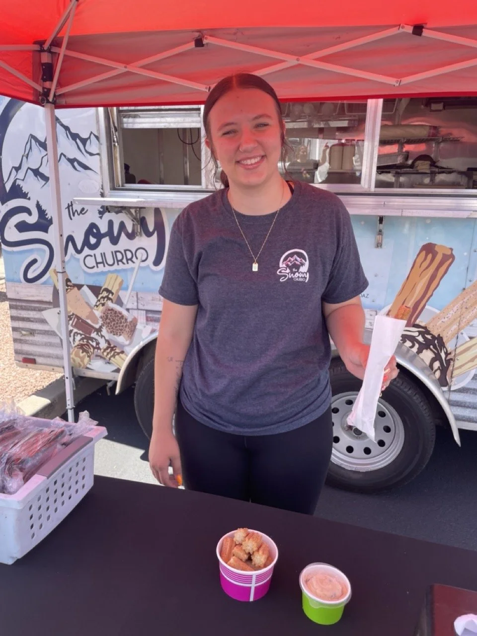 Smiling vendor standing under a red canopy in front of The Snowy Churro food truck, holding a wrapped churro with cups of bite-sized churros and dipping sauce displayed on the table.