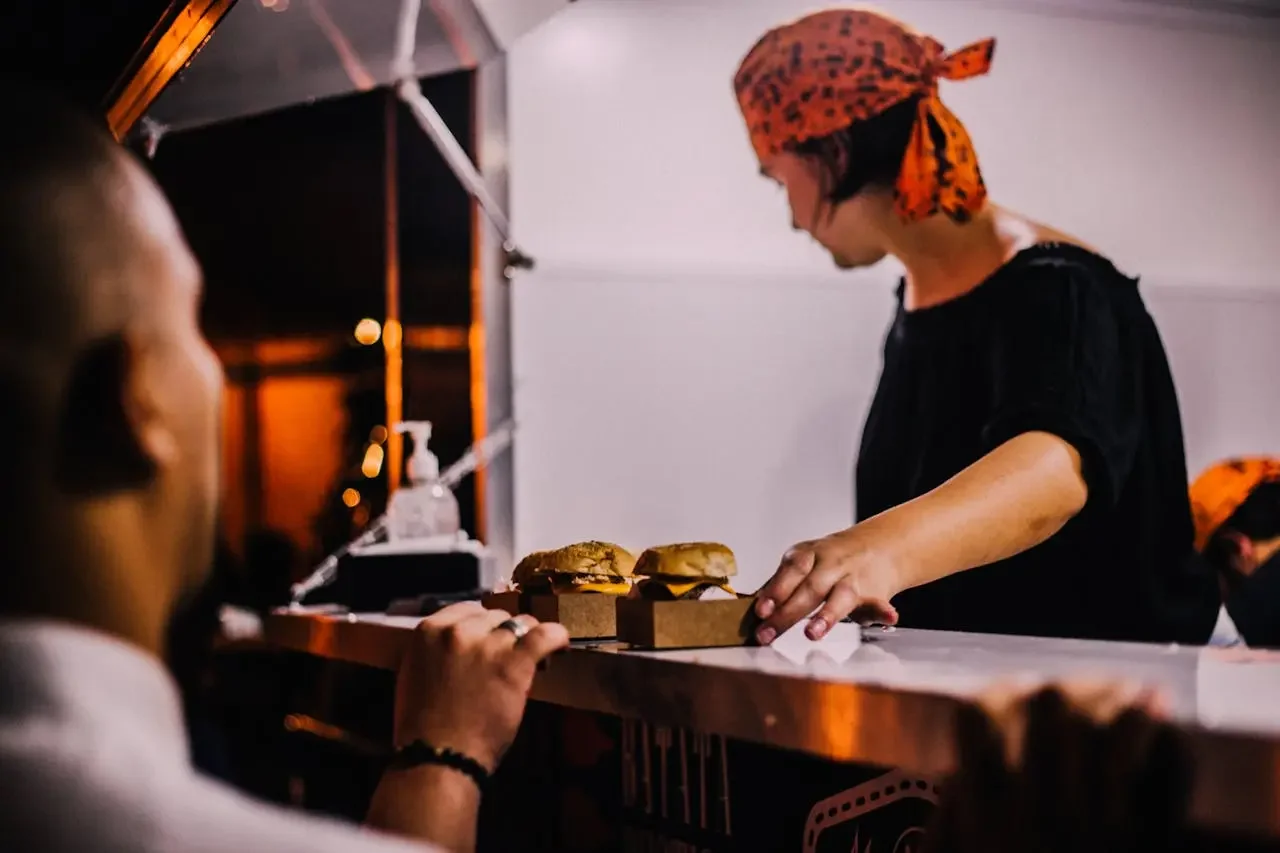 A person wearing an orange bandana serving two hamburgers on a counter at a food truck, with a customer in the foreground.