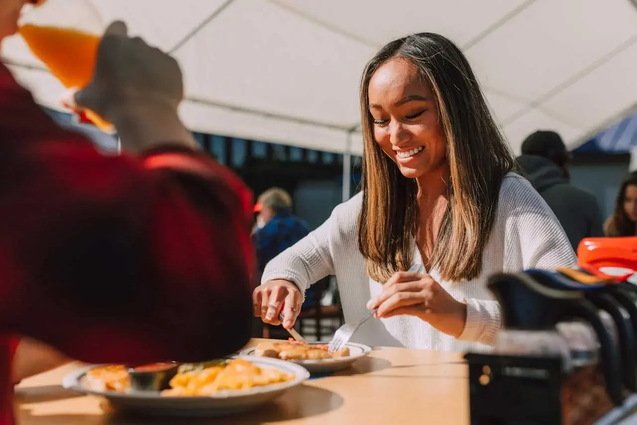 A young woman smiling while eating at a table outdoors under a white open tent, with a plate of food and drinks in front of her. The back of the person seated across from her is in the foreground.